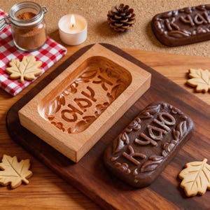 Op de afbeelding: Een rechthoekige houten koekjesvorm met uitgesneden bloemmotieven en cyrillische tekst, vergezeld van een donkerbruin gevormd koekje, lichtgekleurde esdoornbladkoekjes, een glazen pot met bruin poeder, een waxinelichtje en een dennenappel.