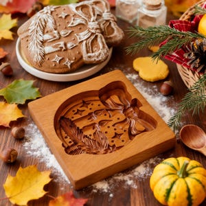 Pu&ograve; includere: Uno stampo per biscotti in legno con un design dettagliato di fiocco, stelle e piume. Un biscotto di pan di zenzero cotto con glassa bianca &egrave; su un piatto. Foglie d'autunno e una piccola zucca sono sparse.