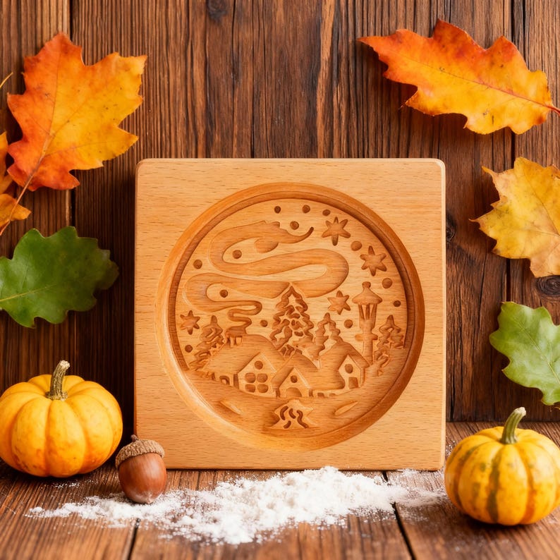 May include: A square, light-colored wooden cookie mold with a detailed winter village scene carved into its surface. The mold is surrounded by fall leaves, pumpkins, and an acorn, suggesting a seasonal theme. The design includes houses, trees, and stars.