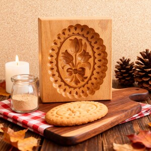 May include: A wooden cookie mold with a floral design is displayed next to a baked cookie on a wooden cutting board. A lit candle, a jar of spices, and pine cones are also in the image. The cookie has a similar floral design.