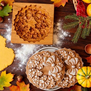 May include: A wooden cookie mold with a wreath design, featuring a bow and holly leaves. Two cookies with the same design are dusted with powdered sugar. The scene includes autumn leaves, a small pumpkin, and a wooden table.