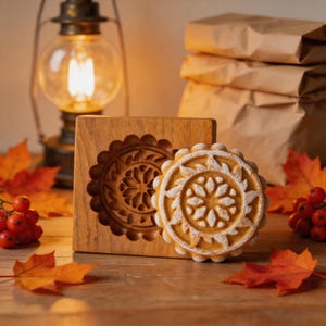 May include: A square wooden cookie mold with a circular floral carving and a matching round cookie featuring a detailed embossed flower pattern, dusted with white powder, positioned together on a wooden surface with autumn leaves.