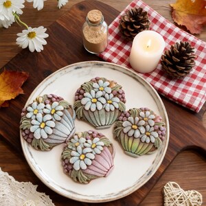 May include: Four flower-shaped cookies on a white plate. Each cookie is decorated with a basket of daisies in shades of blue, yellow, and green. The cookies sit on a wooden board with a candle, pine cones, and a jar of spices.