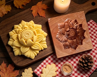 May include: A yellow flower-shaped cookie, a square wooden mold with a carved flower design, two oak leaf-shaped cookies, a small glass jar of light brown powder, and a pinecone arranged on a wooden board and a red and white checkered cloth.