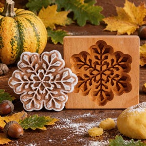 May include: A light brown wooden biscuit mould featuring a carved snowflake design, placed next to a matching snowflake-shaped biscuit with white dusting, a yellow pumpkin, and scattered acorns on a dark surface with flour.
