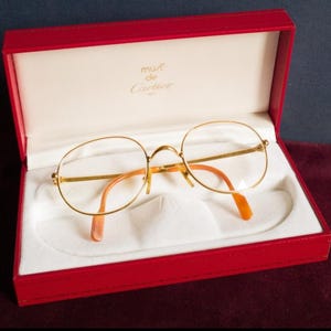 May include: Gold-rimmed spectacles with orange arms sit in an open red box with a white interior. The box displays the words "must de Cartier Paris". The spectacles have a classic, round shape.