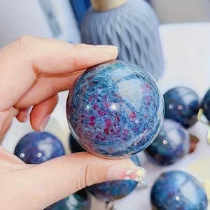 Può includere: Una sfera di pietra preziosa lucidata con un motivo screziato blu e rosso. La sfera è tenuta in mano, con altre sfere simili visibili sullo sfondo. La pietra preziosa ha una finitura lucida.