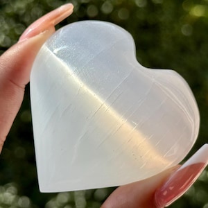 May include: A heart-shaped selenite crystal, a translucent white stone with subtle striations. The crystal is held up against a blurred green background, catching the light and displaying its natural texture. A decorative item.