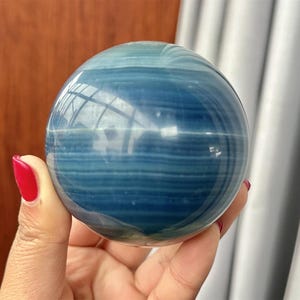 May include: A blue and teal banded, polished sphere. The orb is held in a hand with red nail polish. The surface reflects light, creating a glossy appearance. This decorative sphere is a collectable item.