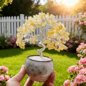 Puede incluir: Una escultura decorativa de árbol con un tronco y ramas de alambre plateado, adornada con piedras de cristal amarillas. El árbol está en una pequeña maceta redonda de cerámica moteada. El fondo presenta una valla blanca, hierba verde y flores rosas.