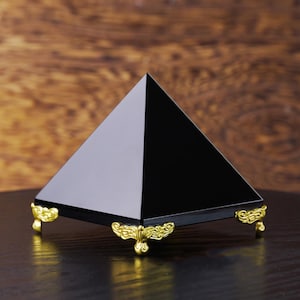 May include: A black pyramid ornament with a shiny surface. It rests on a black base with decorative gold-coloured feet. The background features a wooden surface.
