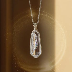 May include: A clear crystal pendant necklace with a silver chain. The crystal is encased in a silver wire frame and has internal rainbow-colored inclusions. The background is a gradient of gold and brown.