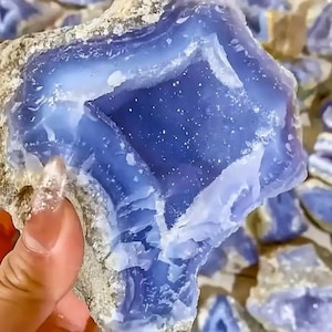 Puede incluir: Primer plano de una piedra preciosa de calcedonia azul con un interior cristalino. La piedra tiene una apariencia en capas con tonos de azul claro y oscuro. El exterior rugoso es de color marrón claro.