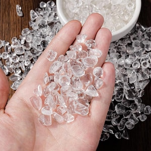 May include: A handful of clear quartz crystal chips, with a scattering of the same crystals on a wooden surface. A small white bowl filled with more crystals is in the background. The crystals are irregular in shape and vary in size.