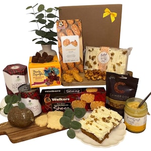 Może przedstawiać: Koszyk prezentowy wypełniony szkockimi przysmakami, w tym herbatnikami, herbatnikami cheddar, fudge i kremem cytrynowym. Brązowy kosz prezentowy ma żółtą kokardę, małą roślinę i kawałek ciasta.