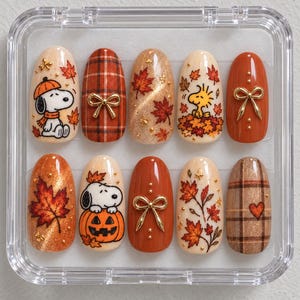 Puede incluir: Un juego de diez uñas postizas en un estuche de plástico transparente, con diseños de temática otoñal que incluyen a Snoopy, Woodstock, patrones de cuadros naranjas, lazos dorados y hojas de arce rojas sobre fondos crema, naranja y dorados.