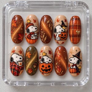 Puede incluir: Un juego de diez uñas postizas ovaladas con diseños de otoño. Las uñas muestran a los personajes Snoopy y Woodstock, calabazas, hojas de arce, patrones de cuadros y esmalte magnético de ojo de gato en tonos marrón, naranja y dorado.
