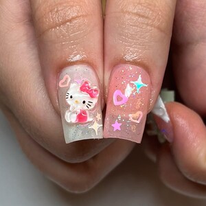 Peut inclure: Gros plan sur deux ongles manucur&eacute;s. L'un pr&eacute;sente un motif Hello Kitty en 3D avec un n&oelig;ud et une robe roses, sur un fond scintillant. L'autre ongle a des paillettes roses avec des c&oelig;urs et des &eacute;toiles.