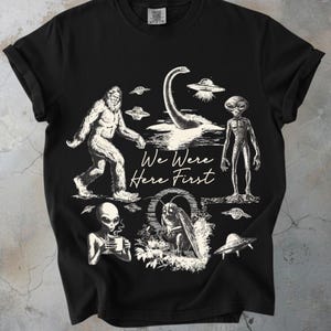 Peut inclure: T-shirt noir avec un motif blanc représentant Bigfoot, le monstre du Loch Ness, des extraterrestres et des ovnis. Le texte "We Were Here First" est imprimé sous les illustrations. Le t-shirt a des manches courtes et un col rond.