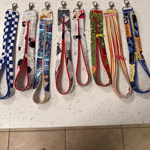 May include: A collection of eight fabric lanyards with various patterns and designs. Each lanyard features a zipper closure and a metal clasp. The patterns include checkered, Minnie Mouse, and other colorful designs. The lanyards are laid out on a light-colored surface.