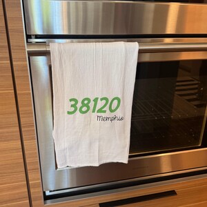 May include: White kitchen towel with the green text "38120 Memphis" hanging on an oven door. The towel is made of a soft, absorbent material, perfect for drying dishes or wiping up spills.