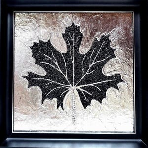 Pode incluir: Obra de arte emoldurada com um desenho de folha preta com veias brancas sobre um fundo prateado texturizado. A folha é preenchida com pequenas partículas escuras, criando contraste com a prata. A obra está contida numa moldura preta.