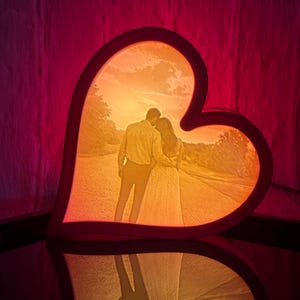 May include: A heart-shaped lamp with a photo of a couple embracing, illuminated with warm orange and yellow light. The lamp has a dark brown frame and is reflected on a dark surface.