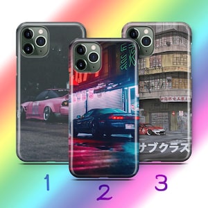 May include: Three smartphone cases featuring automotive designs. Case 1 shows a pink car on a dark street. Case 2 displays a dark car in a neon-lit urban setting. Case 3 depicts a red car parked in front of a multi-story building with Japanese text.