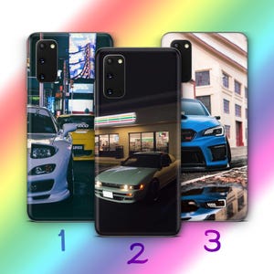 Puede incluir: Tres fundas para teléfono con diseños de coches. La primera funda presenta un coche blanco en un entorno urbano. La segunda funda muestra un coche verde claro frente a una tienda. La tercera funda muestra un coche azul en la calle.