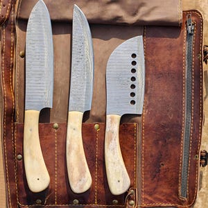 May include: Three stainless steel chef's knives with bone-coloured handles are displayed in a brown leather knife roll. The blades have a patterned design. The knife roll is open, revealing the knives in individual slots.