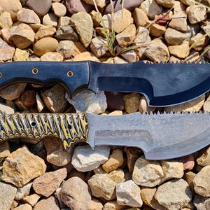 May include: Two custom knives with serrated blades rest on a bed of rocks. The top knife has a dark blue handle with brass rivets and a black blade. The bottom knife features a patterned yellow and black handle and a silver blade.