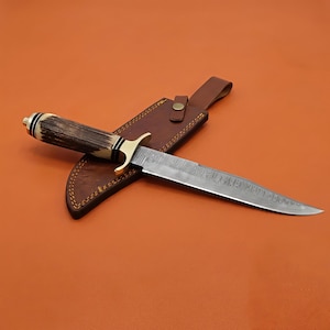 May include: A fixed-blade knife featuring a Damascus steel blade with a visible wavy pattern, a brass guard, and a textured brown antler handle. The knife is accompanied by a brown leather sheath with visible stitching and a brass snap closure.