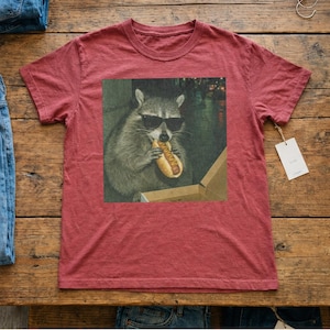 Puede incluir: Camiseta granate con un gr&aacute;fico de un mapache con gafas de sol comiendo un perrito caliente. El mapache est&aacute; en una caja, con un fondo borroso. Una etiqueta est&aacute; adherida a la camiseta.