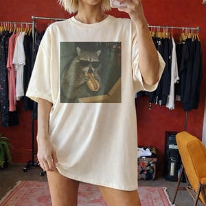 Puede incluir: Camiseta crema de gran tama&ntilde;o con un estampado de un mapache con gafas de sol comiendo un perrito caliente. El gr&aacute;fico est&aacute; centrado en la parte delantera. La camiseta tiene mangas cortas y un corte holgado, adecuada para uso casual.