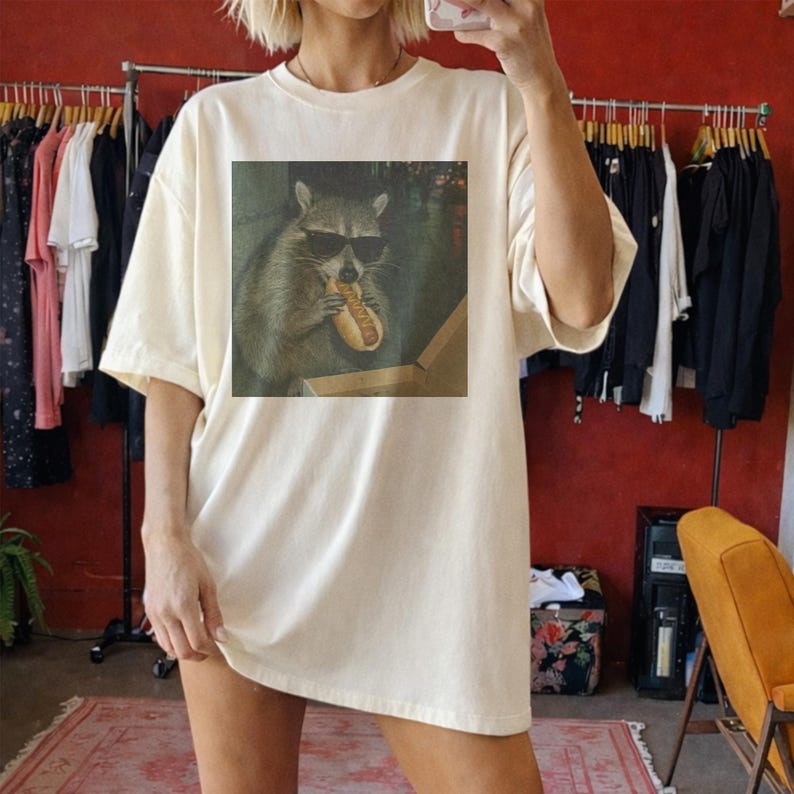 Puede incluir: Camiseta crema de gran tama&ntilde;o con un estampado de un mapache con gafas de sol comiendo un perrito caliente. El gr&aacute;fico est&aacute; centrado en la parte delantera. La camiseta tiene mangas cortas y un corte holgado, adecuada para uso casual.