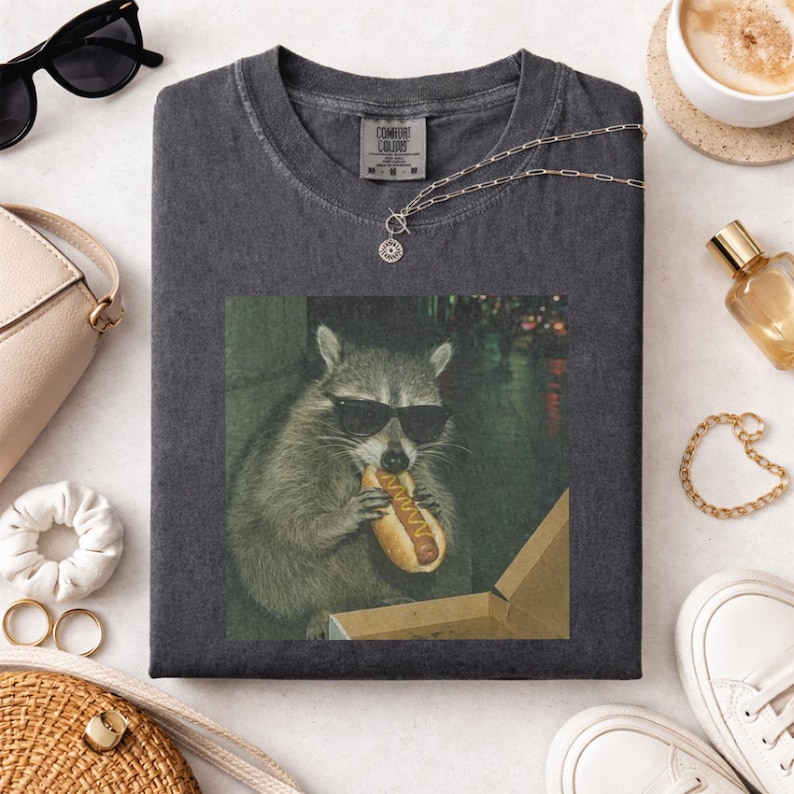 Puede incluir: Camiseta gris oscuro con un dise&ntilde;o de un mapache con gafas de sol comiendo un perrito caliente. La imagen es cuadrada. La camiseta tiene cuello redondo y mangas cortas. La etiqueta de la marca es visible.