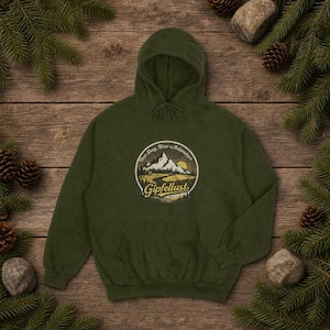 Könnte beinhalten: Ein olivgrüner Kapuzenpullover mit einem kreisförmigen Grafikdesign mit einer Bergkette, Sonne und den Worten "New Day, New Adventure" und "Gipfellust". Der Pullover ist aus weichem Material.