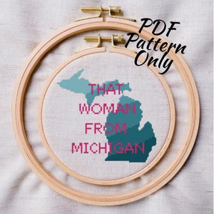 May include: A cross-stitch pattern of the state of Michigan with the text "That Woman From Michigan" in pink. The pattern is in a wooden embroidery hoop.