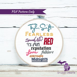 May include: A white embroidery hoop with a cross-stitch design featuring the names of Taylor Swift's albums in different colors. The albums are: Taylor Swift, Fearless, Speak Now, Red, 1989, reputation, Lover, folklore, evermore, and Midnights.