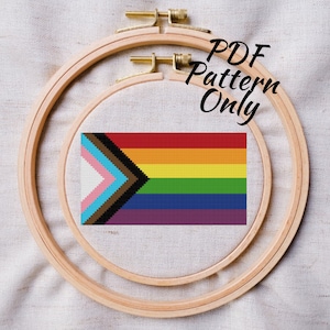May include: A PDF pattern for a cross-stitch project featuring a pride flag with a black and brown chevron on a rainbow background. The flag is displayed in a wooden embroidery hoop.