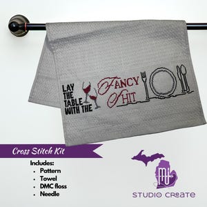 May include: Cross stitch kit with a white towel featuring the text "Lay the table with the fancy shit" and a design of wine glasses, a plate, and silverware. The kit includes a pattern, towel, DMC floss, and needle.