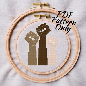 May include: A cross-stitch pattern of three raised fists in different skin tones. The pattern is in a wooden embroidery hoop. The text "PDF Pattern Only" is written in white on a black background.