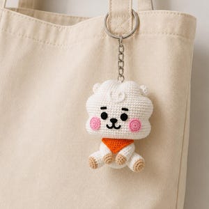 May include: A white crocheted RJ character keychain attached to a beige canvas tote bag. The plush figure features a small orange bandana, pink circular cheeks, black embroidered eyes, and a nose, with light brown accents on the feet and ears.