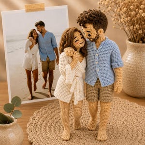Puede incluir: Dos muñecos de ganchillo que representan a una pareja de pie. La muñeca tiene el pelo castaño y viste una bata blanca. El muñeco tiene el pelo castaño, barba y viste una camisa azul claro con pantalones cortos de color tostado. Ambos descalzos.