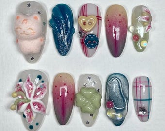 May include: A collection of ten artificial nails with diverse designs. The nails showcase a variety of colours, including pink, blue, and green, and are decorated with 3D embellishments like a cat, flowers, buttons, and glitter. Each nail has a unique design.
