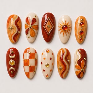 Puede incluir: Un juego de diez uñas postizas en forma de almendra con una paleta de colores cálidos en naranja, crema y rojo oscuro. Los diseños incluyen líneas onduladas, motivos florales, un diamante, un sol, un patrón de cuadros, lunares y fases lunares.