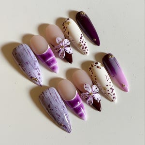 Pode incluir: Uma coleção de dez unhas artificiais longas em forma de amêndoa. Os designs incluem mármore roxo, branco com manchas marrons e roxo degradê. Algumas unhas apresentam pequenos enfeites dourados e flores 3D.