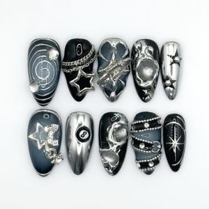 May include: A collection of ten artificial nails with diverse designs. The nails showcase silver, black, and grey hues, adorned with stars, chains, and abstract forms. Some nails have a glossy finish, while others are matte.