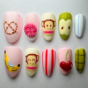 May include: A collection of ten artificial nails, each with a unique design. The designs feature a pretzel, a pink flower, a monkey face, a green apple, blue and white stripes, a banana, red and white stripes, a cherry, and a green grid.