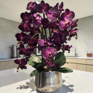 Może przedstawiać: Duża sztuczna orchidea o ciemnofioletowych i purpurowych płatkach, z wieloma rozwiniętymi kwiatami i małymi pąkami. Roślina ma szerokie zielone liście u podstawy i jest umieszczona w okrągłej, metalicznej srebrnej doniczce o teksturowanym wykończeniu.